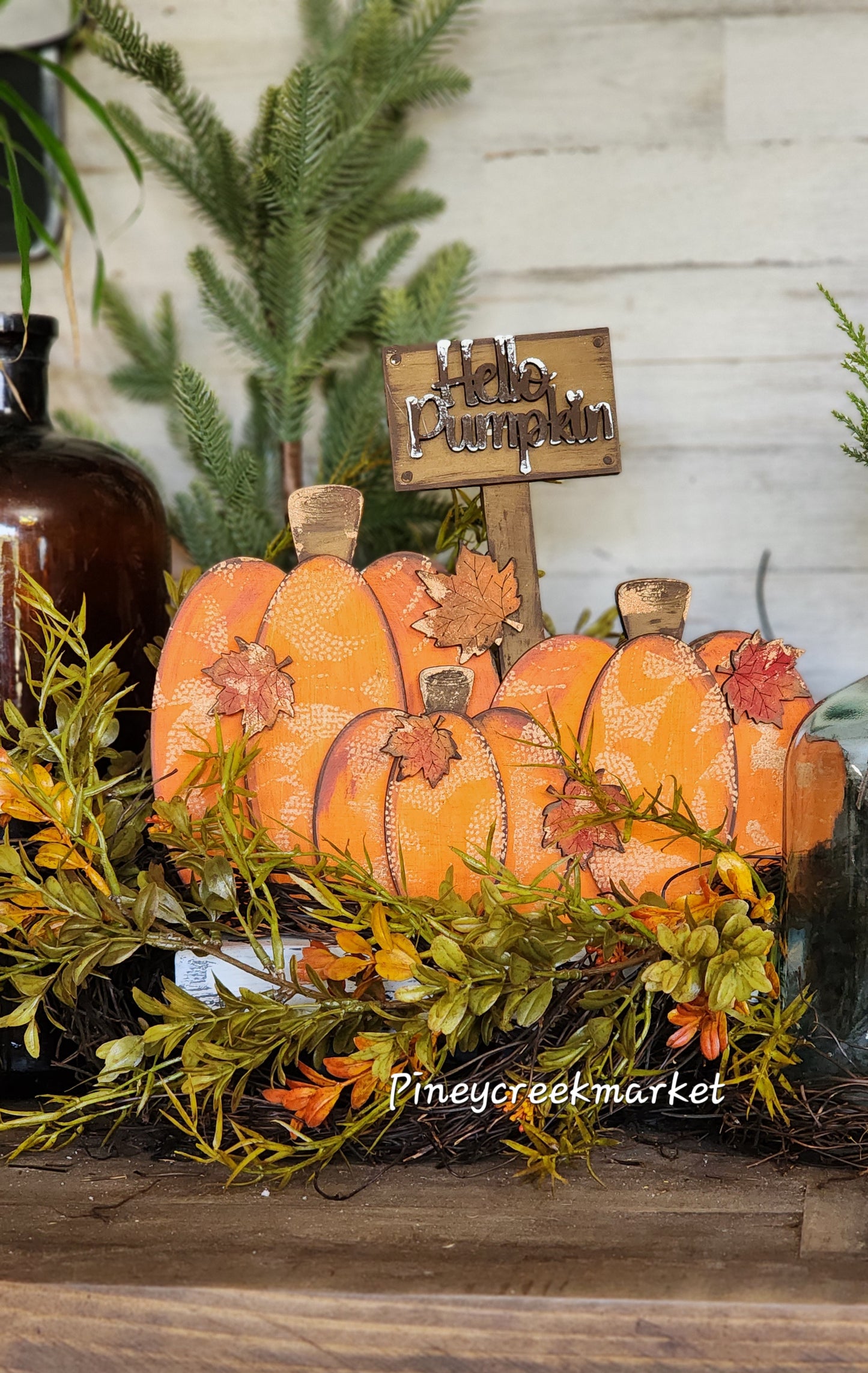 A set of three artificial orange pumpkins with green stems and leaves, and a 'Hello Pumpkin' sign in the background, all placed on a table with autumn-themed decor.